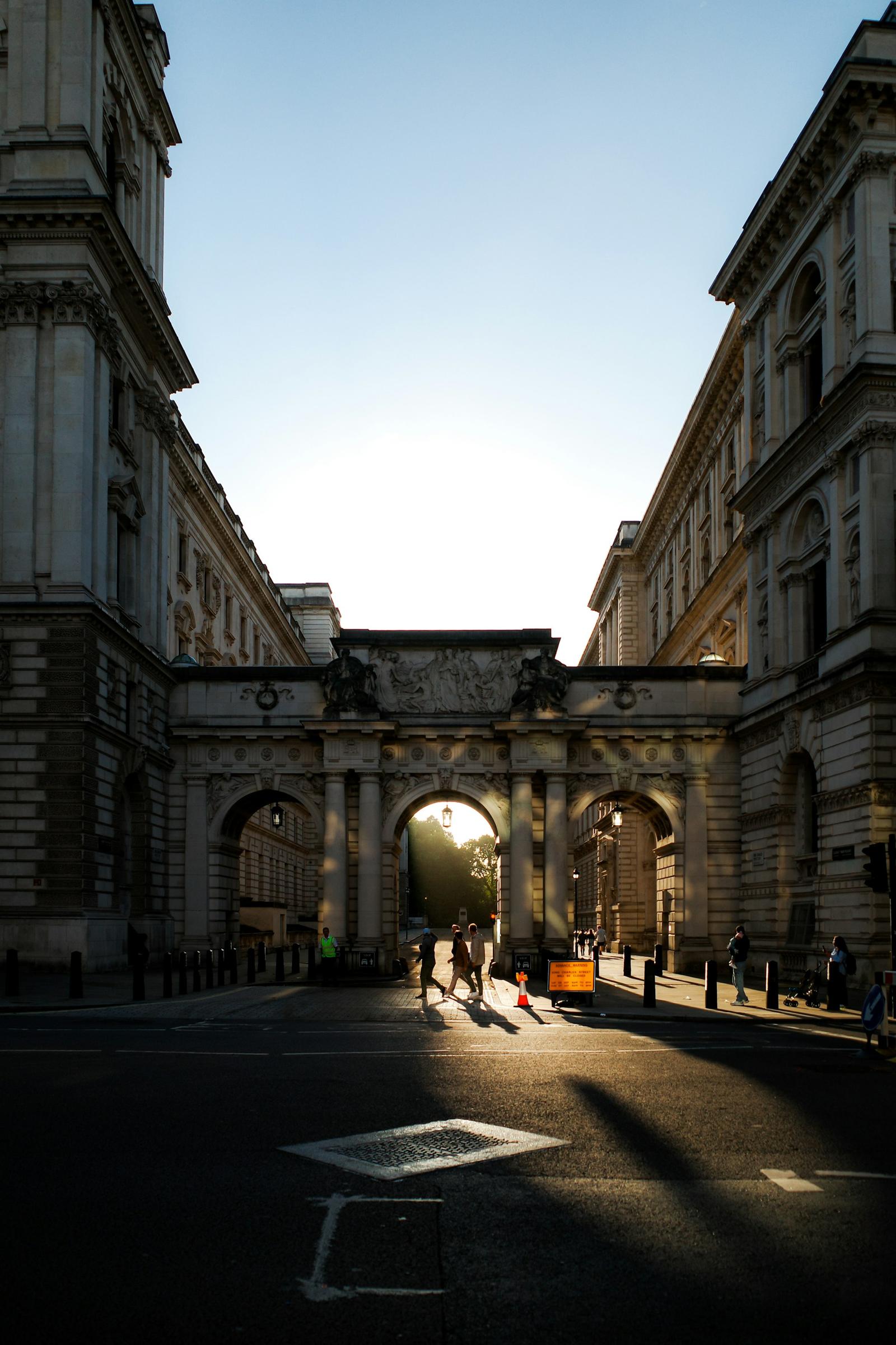 People walking beneath Admiralty Arch in London as low sun streams through the archway.